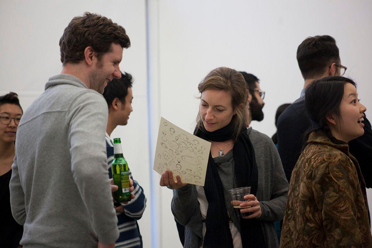 There are various people in a white room. One light-skinned dark blonde or light brown hair woman is looking at the back of a weird faces print. There is a light skinned brown haired man on her left smiling and holding a green bottle.