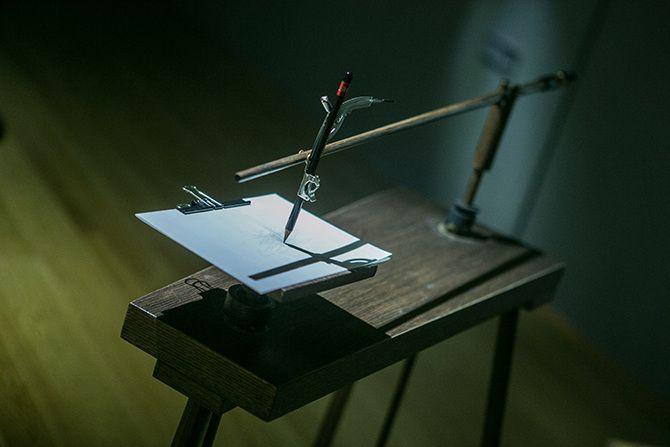 This is a photo of a device that seems to be for drawing. There is a pen and paper pad in the foreground of the picture. The pen is held up by a wooden rod and a metal attachment. The paper pad and wooden tool are supported by a dark block of wood.