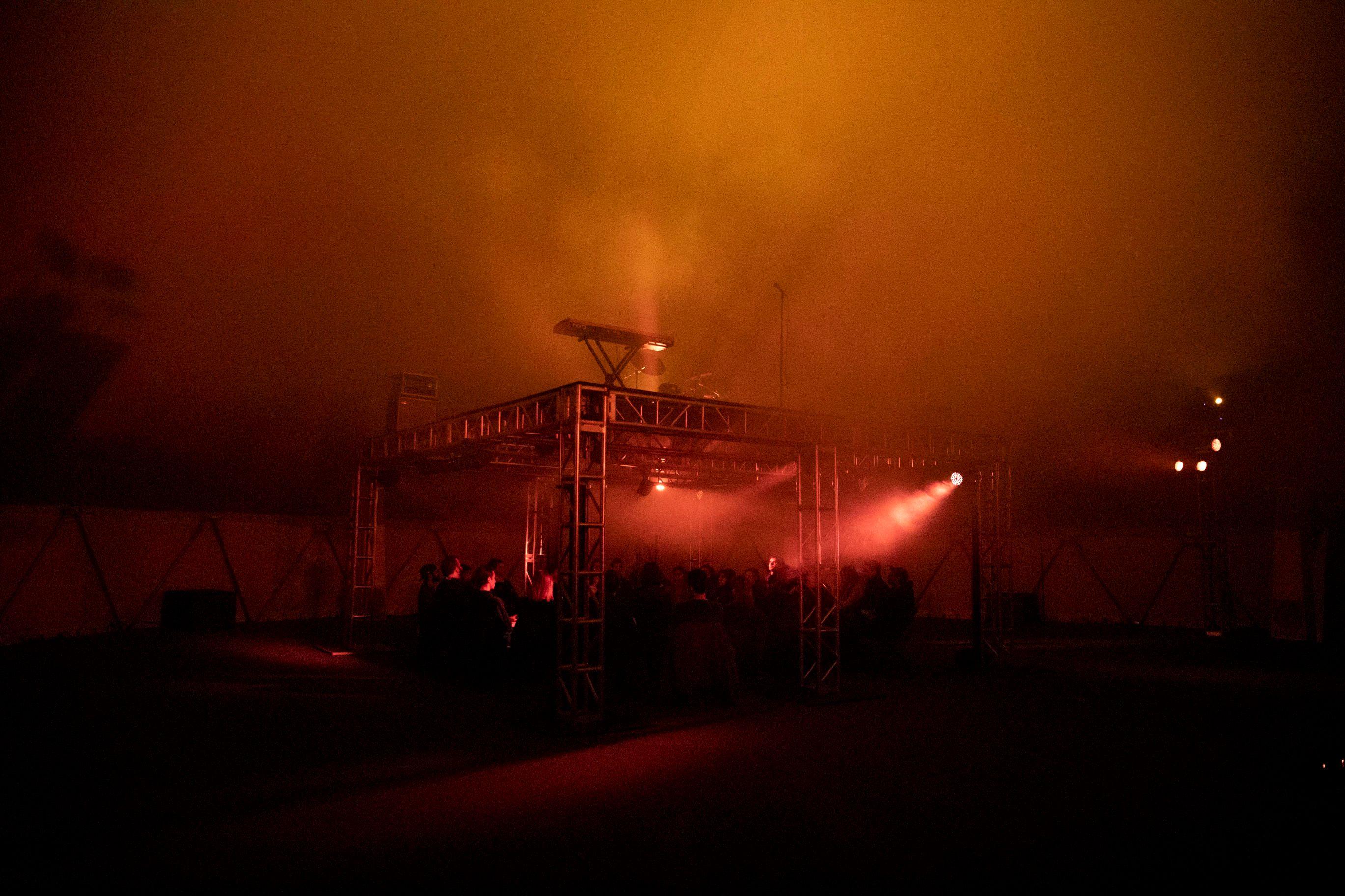 People surrounding a stage setup with lights and scaffolding. There is fog pervading the scene. Red and orange lights make the fog glow.