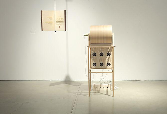 There are two objects in a plain beige room: an oversized book suspended in the air and a machine to its right. The machine has six speakers and one large spinning cylinder with a string attached, all of which are supported by a thin beige wooden structur