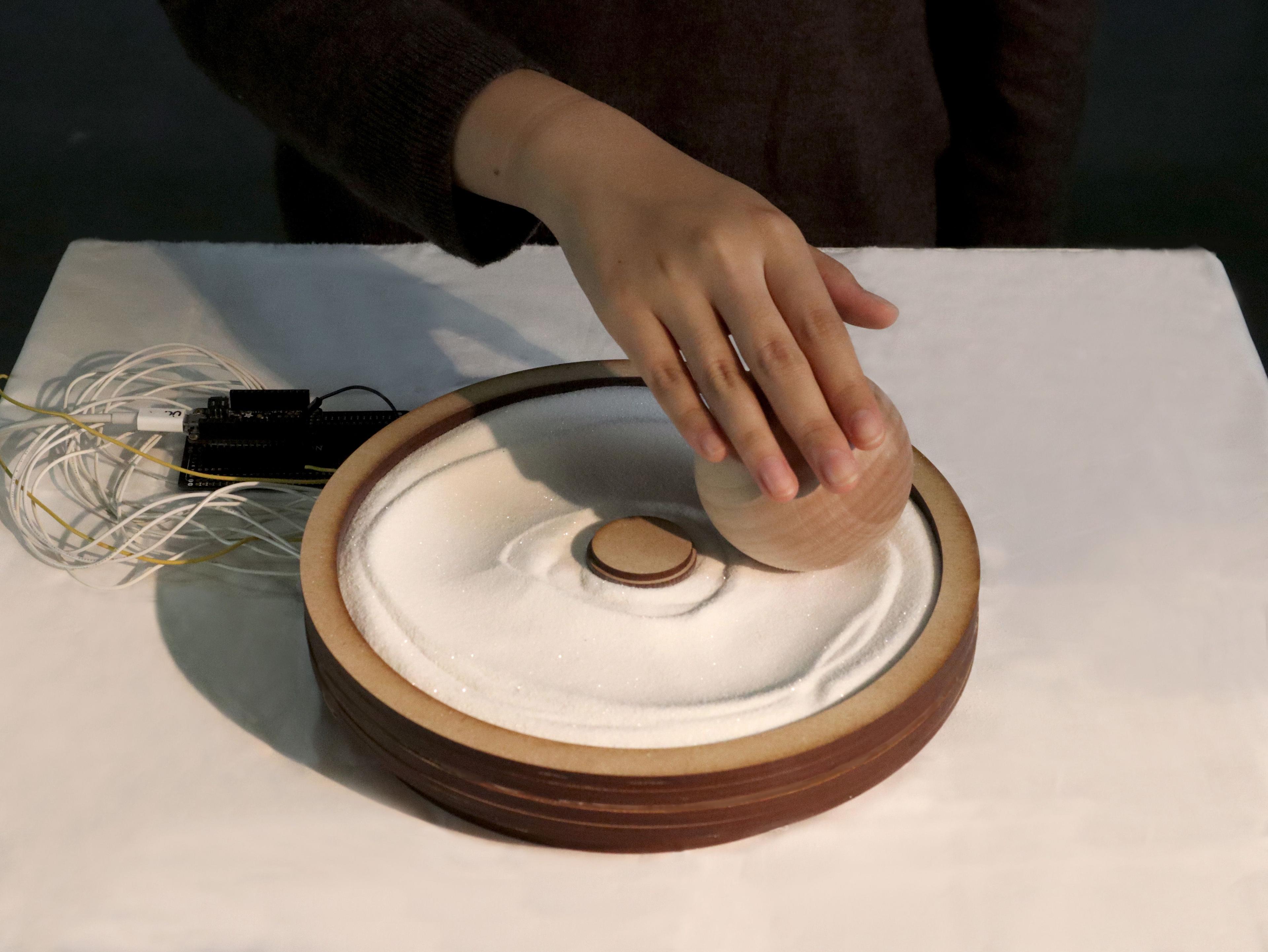 a round container of sand on a table. a hand is combing the sand with a wooden tool. there are wires coming from the sand vessel.