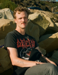 Henrik, a white male in his 30s, with blonde hair and a moustache, sitting on some rocks on a hillside wearing a Deicide shirt